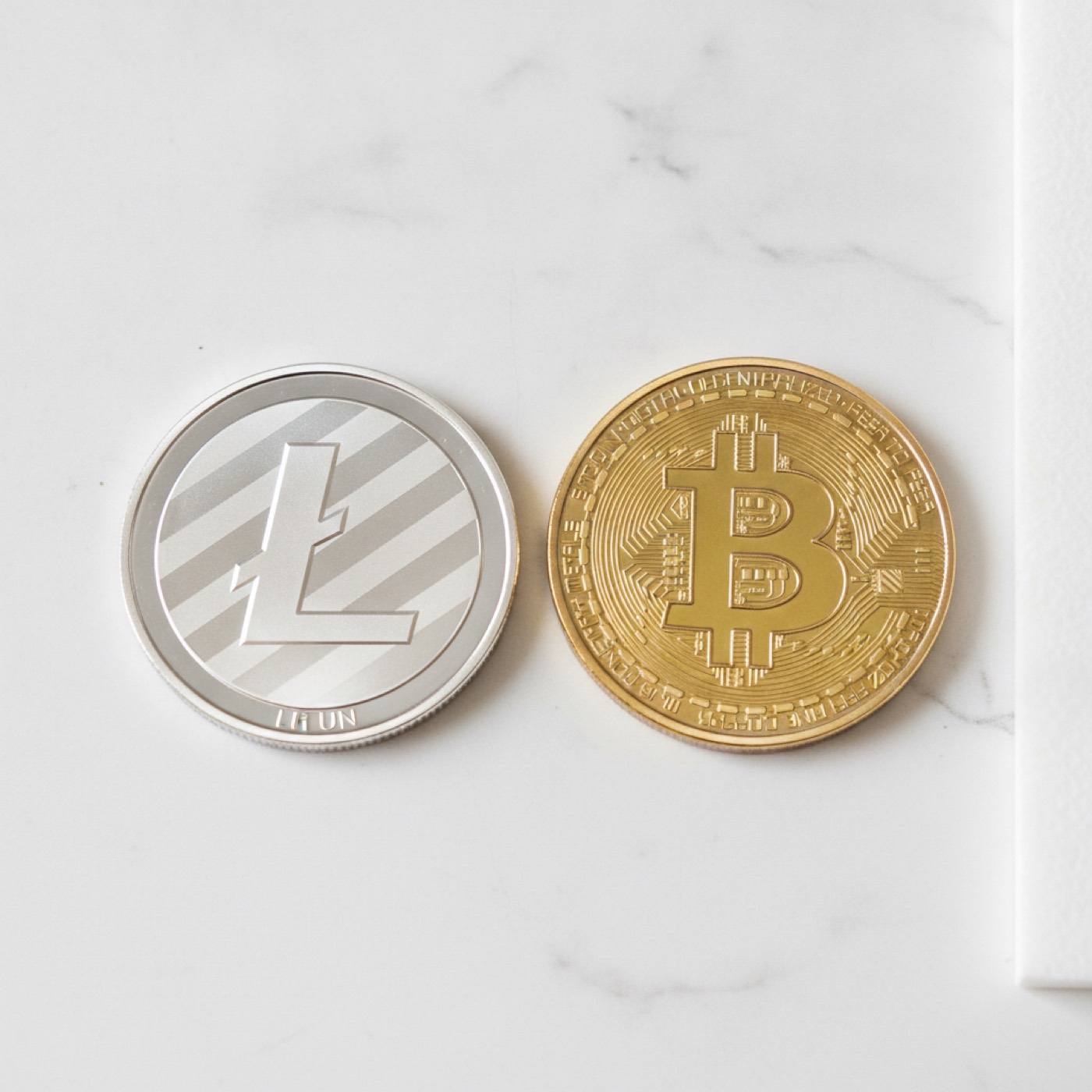 A silver Litecoin coin and a gold Bitcoin coin side by side on white marble